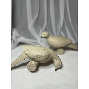 May include: Two off-white decorative bird figurines. One bird is positioned with its head down, while the other stands upright in the background. The birds have a distressed, aged appearance, with visible texture and dark accents on their beaks and eyes.