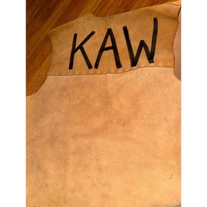 May include: A tan leather piece with the word "KAW" in black block letters. The leather has a rough texture and a natural, earthy tone. The stitching is visible along the top edge.