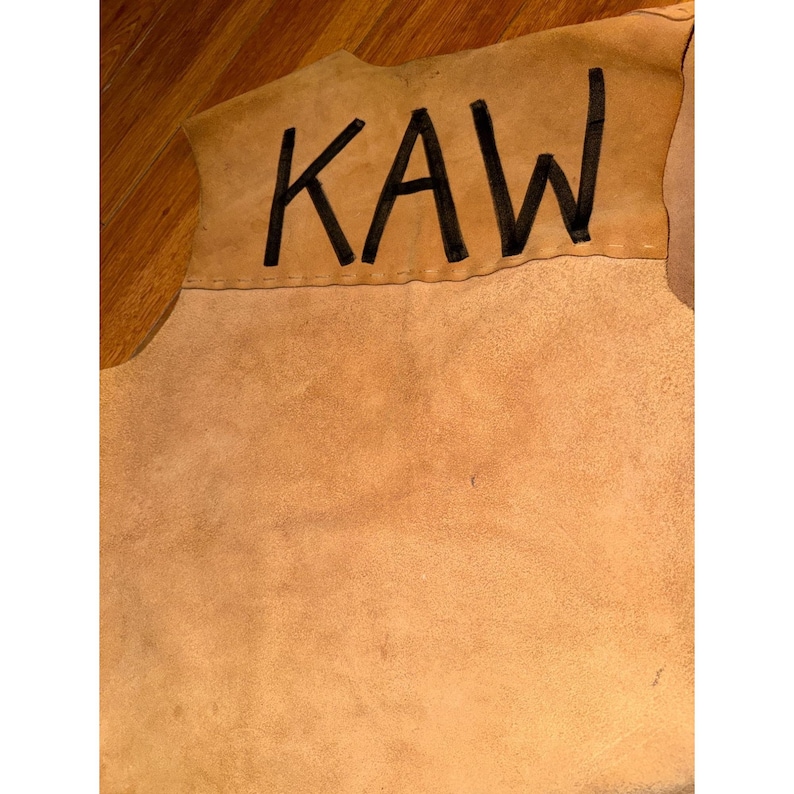 May include: A tan leather piece with the word "KAW" in black block letters. The leather has a rough texture and a natural, earthy tone. The stitching is visible along the top edge.