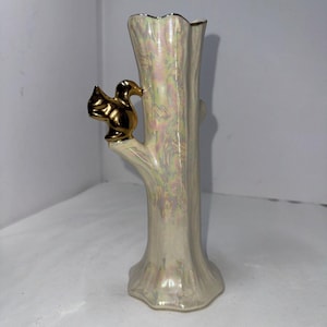 May include: A white ceramic vase shaped like a tree trunk with a gold bird perched on a branch. The vase has an iridescent finish.