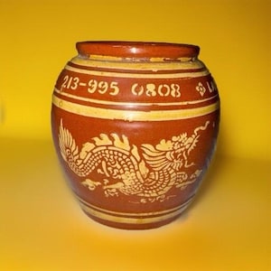 May include: A round, brown ceramic jar with a yellow rim and accents. The jar features a detailed yellow dragon design and the numbers "213-995 0808" in a band around the middle. The jar has a slightly textured surface.