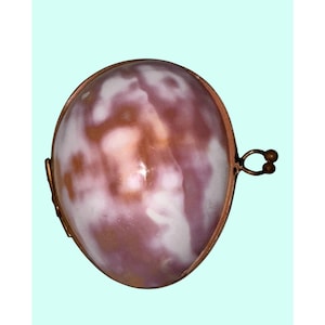 May include: Oval-shaped pendant with a marbled pink and white design, encased in a copper-colored metal frame. The pendant has a small loop at the top for attachment. The background is a light blue.