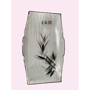 Puede incluir: Bandeja rectangular de cerámica con diseño de bambú. La bandeja tiene un fondo gris claro con un borde dorado. El bambú está representado en negro y dorado. La bandeja tiene texto en la esquina superior derecha.