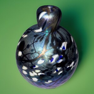 Puede incluir: Un jarrón decorativo de vidrio con un cuerpo bulboso y un cuello estrecho. El jarrón presenta una combinación de colores azul oscuro y negro con acentos blancos y azules. El diseño incluye un patrón de formas y líneas orgánicas.