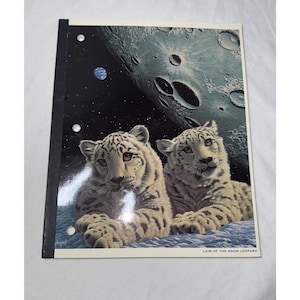 May include: Notebook featuring two snow leopards resting on a blue surface, with a backdrop of a moon and stars. The notebook has a black spine and the text "LAIR OF THE SNOW LEOPARD" at the bottom.