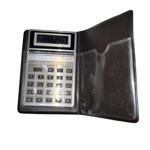 May include: A vintage Casio Mini-Card calculator in a black leatherette case. The calculator has a silver face with black buttons and a digital display. The case is open, revealing the calculator and a pocket.