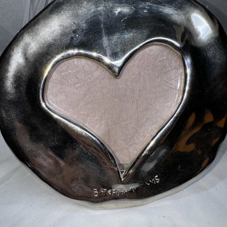 May include: A silver-colored, heart-shaped decorative plate with a textured surface. The center features a heart-shaped cutout with a light pink fabric insert. The words "Barefoot Tums" are etched at the bottom.