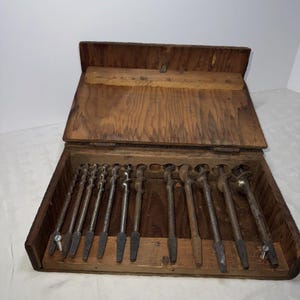 May include: A wooden box with a hinged lid containing a set of 12 drill bits. The box is open and the drill bits are visible in their slots.