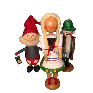 May include: Three wooden figurines: a gnome in a red hat, a woman in a traditional dress, and a soldier with a green hat. The gnome has a black body with red braces. The woman has blonde hair and a white dress. The soldier has a white beard.