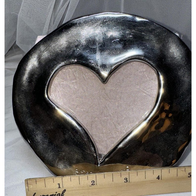 May include: A silver-toned picture frame with a heart-shaped opening. The frame has a metallic finish and a soft, neutral-colored backing. The frame is approximately 4 inches wide.