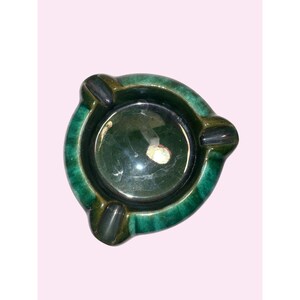May include: A round ceramic ashtray featuring a deep forest green glaze with darker accents. The piece has a smooth, glossy finish and includes three distinct notches along the rim for resting items, surrounding a central circular basin.