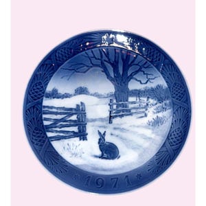 May include: A round blue and white porcelain decorative plate featuring a winter landscape with a rabbit in the foreground, a wooden fence, and a large tree. The border has a relief pattern of pinecones, and the year 1971 is printed at the bottom.