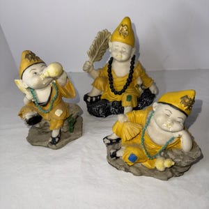 Könnte beinhalten: Drei kleine, cremefarbene Mönchsfiguren in gelben Roben und Hüten. Einer trinkt aus einer Kalebasse, ein anderer hält eine Feder und der dritte ruht sich aus. Jede Figur hat eine Perlenkette und sitzt auf einem Felsensockel.