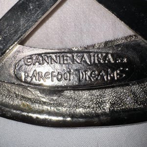 May include: Close-up of a silver-toned metal object with the text "ANNIE KAJIKA" and "BAREFOOT DREAMS" embossed on it. The object has a textured surface and a curved shape.