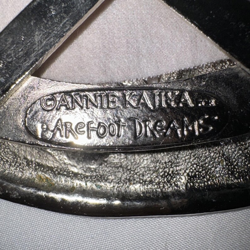 May include: Close-up of a silver-toned metal object with the text "ANNIE KAJIKA" and "BAREFOOT DREAMS" embossed on it. The object has a textured surface and a curved shape.