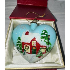 Könnte beinhalten: Herzförmiger Weihnachtsschmuck mit einer Winterszene. Der Schmuck zeigt ein rotes Haus mit weißem, schneebedecktem Dach, grüne Bäume und einen Schneemann. Der Hintergrund ist hellblau. Der Schmuck befindet sich in einer roten Schachtel.