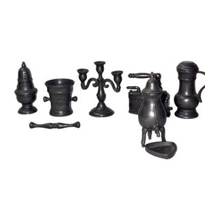 May include: A collection of miniature pewter-colored metal items including a shaker, a mortar and pestle, a three-armed candelabra, a small iron, a coffee grinder with a handle, and a lidded pitcher, all arranged against a plain white background.