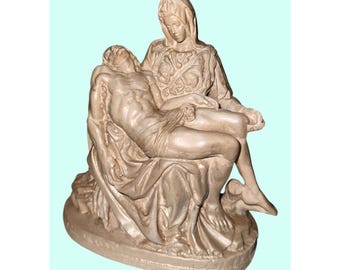 May include: A beige sculpture depicting a seated woman holding a man. The man is lying across her lap. The sculpture is on a round base and is a religious themed item.