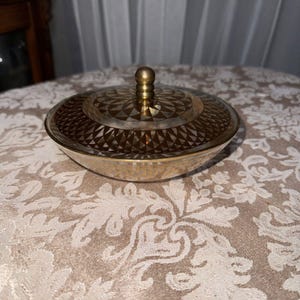 May include: A vintage, gold-toned glass trinket dish with a matching lid. The lid features a geometric pattern and a small, round knob. The dish is round with a slightly flared rim. The overall design is elegant and decorative.