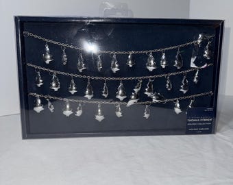 May include: A silver holiday garland with small, tree-shaped ornaments. The garland is displayed in a black box. The text on the box reads "THOMAS O'BRIEN HOLIDAY COLLECTION HOLIDAY GARLAND".