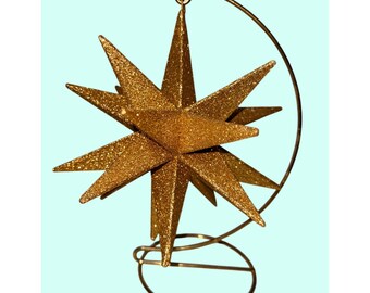 May include: A gold glitter star ornament with multiple points, suspended from a gold metal stand. The star has a textured surface and is designed for decorative purposes. The stand has a circular base and a curved top.