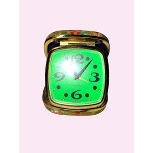 May include: A vintage travel alarm clock with a bright green face and gold-tone metal frame. The clock face features black numerals and hands, with the word "ALARM" visible. The clock is encased in a floral patterned case.