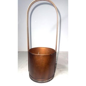 May include: A cylindrical copper-colored metal container featuring a tall, thin, arched handle that extends vertically from the sides. The vessel has a smooth, metallic finish with a circular base and an open top, presenting a simple, functional design.