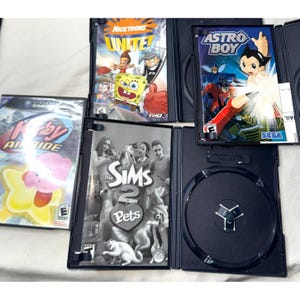 May include: A collection of four video game cases including Kirby Air Ride, Nicktoons Unite!, Astro Boy, and The Sims 2 Pets. The cases are black plastic with colorful cover art featuring various animated characters and game titles printed on the front.