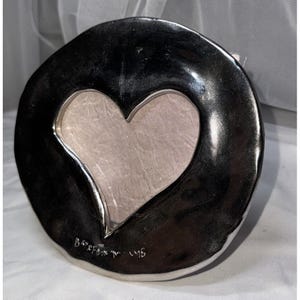May include: A silver-colored, heart-shaped picture frame. The frame has a dark, glossy finish with a heart-shaped cutout in the center. The back of the frame is a light gray color. The frame is marked "Barefoot in the sand" and "96".