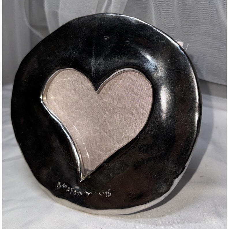 May include: A silver-colored, heart-shaped picture frame. The frame has a dark, glossy finish with a heart-shaped cutout in the center. The back of the frame is a light gray color. The frame is marked "Barefoot in the sand" and "96".