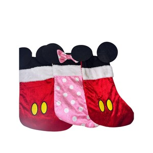 Op de afbeelding: Drie kerstsokken met Mickey Mouse en Minnie Mouse thema's. Twee rode sokken met gele knoopaccenten en zwarte oren. De middelste sok is roze met witte stippen en een roze strik. Allemaal met witte imitatiebont afwerking.