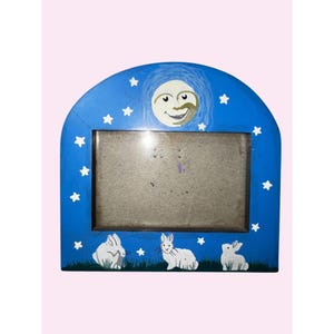 May include: A blue wooden picture frame with a whimsical design. The frame features a smiling moon, white stars, and three white rabbits on a green grassy base. The frame is designed to hold a rectangular photo.