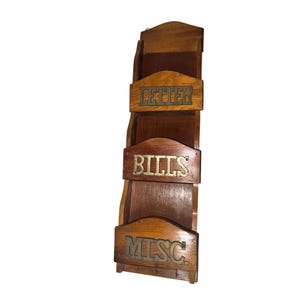 Könnte beinhalten: Ein Vintage-Wandorganizer aus Holz mit drei Fächern, die mit goldfarbenen Buchstaben mit „LETTER“, „BILLS“ und „MISC“ beschriftet sind. Der Organizer ist aus dunklem Holz gefertigt und hat eine gewölbte Oberseite. Er verfügt unten über einen kleinen Schlüsselhalter.