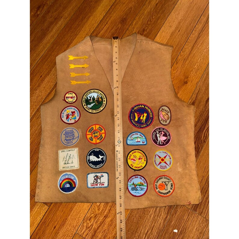 May include: A tan vest with multiple colorful patches, including arrows, circles, and rectangular shapes. The patches feature various designs, such as a whale, a rainbow, and text. A wooden ruler is placed vertically along the vest, measuring approximately 29 inches.