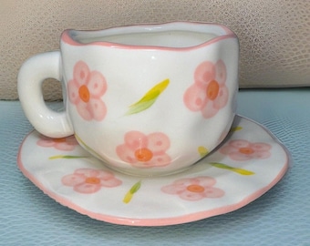 May include: A white teacup and saucer set with a pink floral design. The cup has a scalloped rim and the saucer has a pink trim.