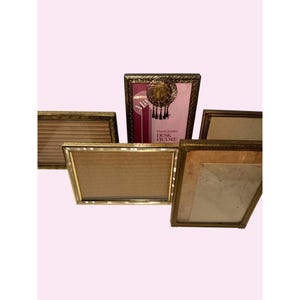 Puede incluir: Una colección de cinco marcos de escritorio rectangulares con bordes dorados metálicos y bordes texturizados. Un marco presenta un inserto rosa con el texto "Finest Quality DESK FRAME" y un emblema decorativo circular con borlas negras colgantes.