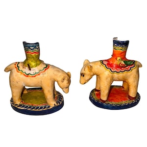 Puede incluir: Dos figuras de oso pintadas a mano. Los osos son de color crema con detalles coloridos en la espalda y las bases. Cada oso tiene una abertura cilíndrica en la espalda, lo que sugiere que pueden ser candelabros o artículos decorativos.