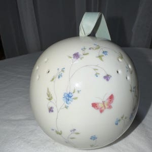 May include: A white, spherical decorative object with a pale blue ribbon. The sphere is decorated with floral and butterfly designs in blue, purple, and pink. Small holes are evenly spaced around the sphere.