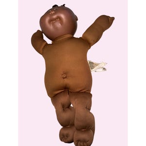 May include: A brown Cabbage Patch Kid doll with a dark brown head and a brown fabric body. The doll has its arms outstretched and its legs bent. A tag is attached to the doll's back.