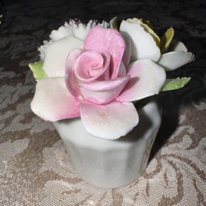 May include: A porcelain figurine of a bouquet of flowers in a white pot. The bouquet features a pink rose with white petals, white flowers, and green leaves.