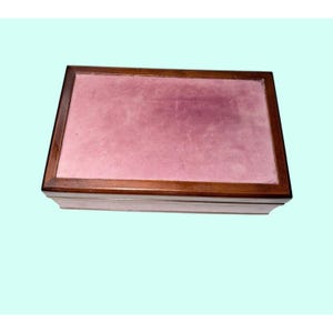 May include: A rectangular wooden box featuring a dark brown polished frame and a flat, soft-textured mauve-pink top panel. The box has a clean, simple design with a smooth finish on the wooden edges and a contrasting matte fabric surface.
