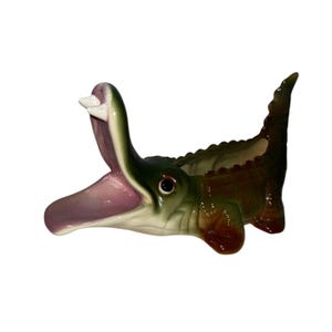 Puede incluir: Una maceta de cerámica con forma de cocodrilo con la boca abierta, mostrando un interior rosa y un solo diente blanco. El cuerpo del cocodrilo es una mezcla de verde y marrón, con un diseño escamoso texturizado. La maceta está diseñada para plantas pequeñas o artículos decorativos.