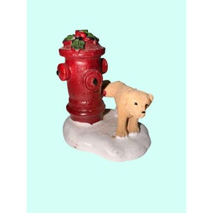 May include: A miniature Christmas figurine featuring a red fire hydrant with holly and berries on top. A small, tan-colored dog stands next to the hydrant on a snowy base. The figurine is likely made of ceramic or resin.
