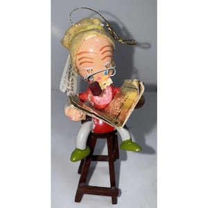 May include: A vintage Christmas ornament of a figure reading a newspaper, seated on a small wooden stool. The figure wears glasses, a hat, and a red shirt. The ornament has a gold hanging loop.