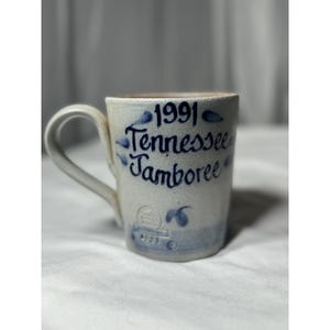 Puede incluir: Taza de cerámica crema con la inscripción azul "1991 Tennessee Jamboree". La taza tiene un asa curva y detalles decorativos azules.