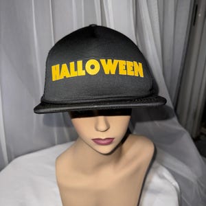 May include: Black trucker hat with the word "HALLOWEEN" in bright yellow block letters across the front. The hat has a curved brim and a mesh back. A fun accessory for Halloween.