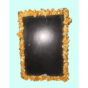 May include: A rectangular mirror with a black surface, framed by a gold-colored, ornate border of roses and leaves. The frame has a textured, three-dimensional appearance, and the mirror is set against a light blue background.