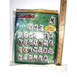 May include: A Bucilla counted cross stitch kit for 30 tiny stocking ornaments in a plastic package, featuring various holiday designs. A wooden ruler is placed vertically next to the package, showing a height of approximately 13.5 inches.