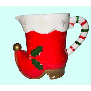 May include: A festive red and white ceramic pitcher shaped like an elf's boot. The pitcher features a gold toe, a white rim with gold star accents, and a handle with red and green stripes. Green holly leaves decorate the side.