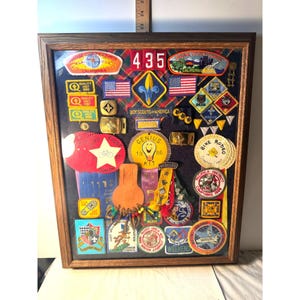 May include: A rectangular wooden frame containing a collection of Boy Scouts of America patches, pins, and badges. The display features various embroidered emblems, including unit numbers 435, American flags, and circular event patches.